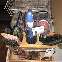 Photo showing multiple hairbrushes including paddle brushes, a vented pink brush, BabyBliss Pro brushes with blue and white handles, held in wooden holders and wire baskets.