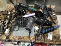 Photo showing tangled hair dryers, diffuser attachments, hair brushes, a hair straightener, and cords on a kitchen countertop.