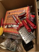 Top view of box showing assorted kitchen utensils with red handles, including cheese grater, juicer, can opener, whisk, and KFC oven mitt set.