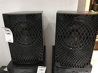 Pair of KEF speakers shown side by side with black protective grills, each tagged with inventory labels.