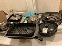 Photo showing a collection of pots, frying pans, baking pans including a black roasting pan with rack, blue baking pan, and portable electric skillet with lid.