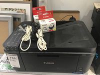Canon Pixma MX492 printer showing front and top with scanner lid and cables on top, with visible packaging box and two Canon ink cartridge boxes behind it.