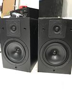 Front view of two black KEF Reference Series SP3079 speakers showing the midrange/tweeter and woofer drivers.