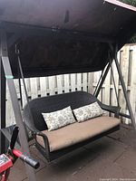 Front view of outdoor swing with canopy, showing brown wicker bench, beige seat cushion, two floral-patterned rectangular pillows, and stained canopy fabric above.