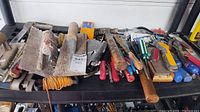 Overview of various hand tools arranged on a black shelf, including trowels, hammer, screwdrivers, wrenches, wire cutters, and other miscellaneous hand tools showing wear.