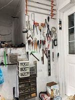 Wall with various hand tools hanging, including saws, clamps, pliers, screwdrivers, hammers, scissors, wrench, and measuring tools.
