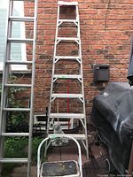 7-foot aluminum ladder and small step ladder visible outdoors against brick wall with garden tools and grill nearby.