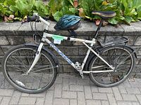 Full side view of white Avalanche touring bicycle with black rear rack and attached water bottle holder. Dark green Bell helmet resting on top tube. Shown on paved surface with stone wall and plants in background.