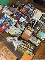 Overhead photo showing a large variety of art supplies spread on a wooden surface including crayons, colored pencils, markers, art stamps, pens, and paint sets in original packaging and plastic bags.