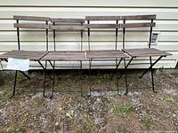 Four folding chairs shown unfolded, featuring black metal frame and multiple light brown wooden slats for seat and backrest, placed outside against a beige wall.