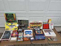 Lot of boxed games and puzzles including party games, trivia, card games, and jigsaw puzzles laid out on garage floor in front of white garage door.