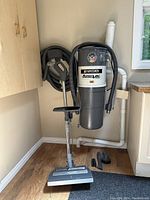 Front view of wall-mounted central vacuum unit with hose coiled and vacuum power head attachment on the floor.