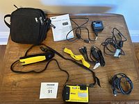 Overview of Nikon Coolpix AW130 camera with accessories laid out on wooden table including carrying bag, manual, charger, wrist straps, and cables.
