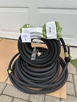 Photo of two coiled black porous soaker hoses with brass fittings, one fitting shown closely with mesh screen inside. Package of galvanized wire hooks labeled to hold hose in place.