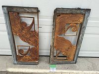 Two rusty iron decorative inserts in wooden frames placed on concrete, showing the scroll and wave design with heavy rust and a weathered wood frame.