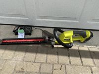 Full length view of Ryobi cordless hedge trimmer with attached 18V battery and charger beside it on pavement.