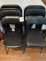 Seven black folding chairs with vinyl padded seats and backrests, stacked and arranged side by side on a wooden floor.
