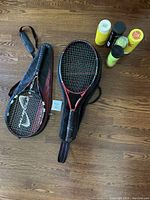 Two tennis rackets side by side, one in a black case with a white swoosh logo, the other resting on the floor, showing red and black colors. Five cans of tennis balls grouped nearby.
