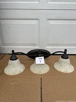 Full view of three globe wall sconce showing milky glass bell-shaped globes and dark metal frame against garage door background.