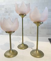 Set of three vintage brass candle holders with blush pink glass shades shaped like flower petals arranged on a white surface against white tile background.