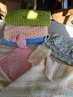 Photo showing stacked hand knitted baby blankets in green, peach, blue, and multicolored yarns along with a pink baby hat with animal ear details and a cream knitted baby bonnet.