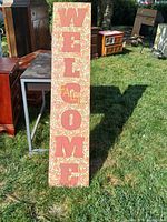 Tall wooden 'WELCOME' porch sign placed on grass showing floral background and red letters