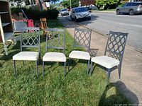Four blue metal framed chairs with white padded seats arranged outdoors on grass next to a sidewalk and road, showing front and angled views.