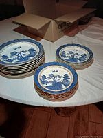Full set of porcelain plates stacked on a table, showing blue and white Willow transferware pattern with gold trim.