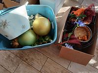 Front view of plastic container holding large glittery green and gold ornaments, small striped ornaments, and artificial white flowers with a box containing floral wire frames, brown mesh ribbon, purple artificial flowers, and small wooden decorative items nearby.
