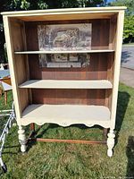 Front-facing image of the decorative wooden display hutch outdoors, showing two white shelves inside, scalloped edge bottom, turned legs, and open front with no doors.