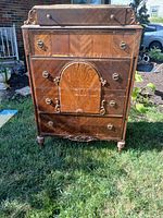 Front view of the antique wooden dresser showing ornate wood paneling, multiple drawers, and metal ring handles. Visible wear on wood surface, especially on the drawers.