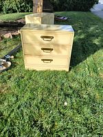 Photo of one cream-colored wooden dresser featuring three drawers with brass handles, placed on grass in daylight.