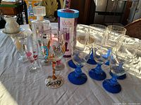 Top down view of glassware on table showing clear glasses with blue stems, decorative stemmed glasses with painted designs and a cylindrical glass bottle with lid inside a cardboard tube.