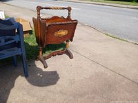 Wooden magazine rack with decorative inlay on the side panel, carved legs, and a handle across the top, placed outdoors on a sidewalk.
