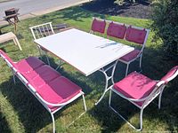 Rectangular white wood top patio table with metal legs, surrounded by red cushioned chairs and benches on grass.