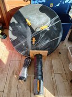 Round black table top, face down, with two detached legs wrapped separately in plastic. Visible damage and wear on legs and table surface.