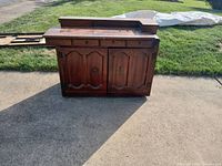Front view of antique wooden sewing cabinet showing two paneled doors and metal handles. Cabinet placed outdoors on pavement.