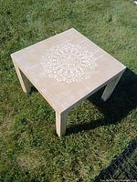 Square wooden side table shown from above on grass with white mandala pattern in the center