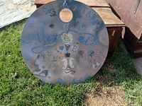Round metal wall shelf with colorful painted artwork, small inset round mirror, and attached hooks for hanging items, placed outdoors on grass.