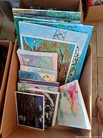 Box containing multiple wall paintings and canvases of various sizes and artistic styles, including nature-themed and abstract images.