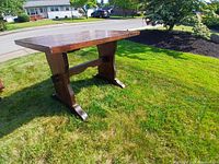 Wooden table shown outside on grass from a side angle, displaying the pedestal legs and stretcher support under the top.
