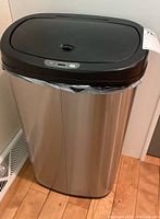 Front angle view of the stainless steel automatic lift top trashcan showing the black plastic lid, control panel, and plastic trash bag inside.