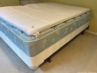 Photo showing queen size mattress with light blue floral pattern on box spring and white mattress topper covering it.