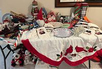 Table display of assorted Christmas holiday decor items including embroidered tablecloth with cardinals, fabric mittens with holiday themes, decorative plate, and plush stuffed animals.