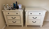 Two white bedside tables side by side, each with three drawers, black knobs and handles on drawers, minor wear visible on edges.