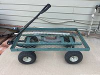 Green metal garden cart with black handle and mesh bottom, four large thick pneumatic tires on a concrete surface outdoors