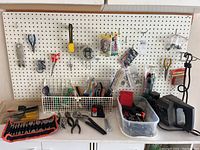 Full view of a pegboard with various small hand tools hanging and laying beneath, including a roll-up tool pouch, pliers, screwdrivers, and a plastic bin with tools.