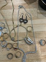 Mixed jewelry items arranged on wooden surface including rings, a silver-tone rhinestone necklace, a gold-tone chain necklace, and heart-shaped earrings.