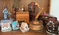 Overview of nine music boxes displayed on a shelf including ceramic miniature houses, wooden harp, cabinet, piano, roll-top desk, and figurines