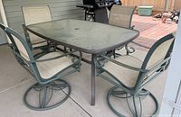 Rectangular patio table with glass top and four metal frame swivel chairs with beige mesh seating around it on concrete patio.